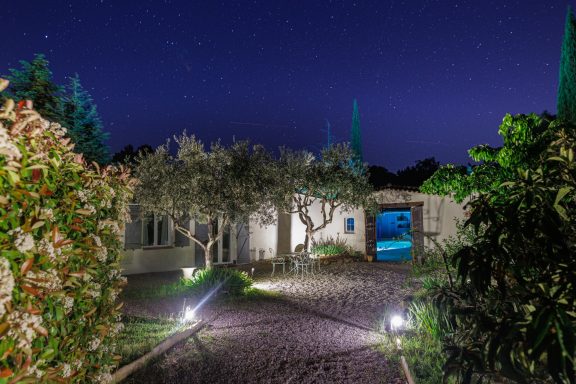 Jardin illuminé de nuit – Suite Mooréa - Var - Chambre d'hôtes Jardin avec arbres, chemin éclairé et porte menant à une piscine sous un ciel nocturne.