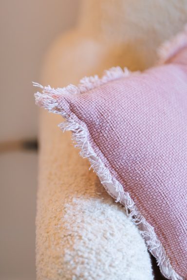 Coussin rose avec des bords effilochés posé sur un canapé texturé.